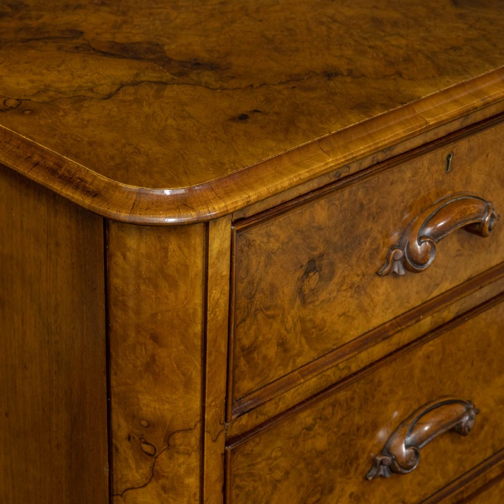 A Victorian burr walnut chest of drawers featuring a two-over-three drawer arrangement with carved handles and rounded corners on a plinth base. View XII