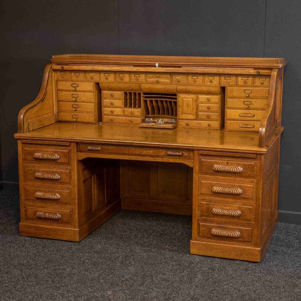 A large Edwardian golden oak roll-top desk featuring a slatted tambour cover, an extensive fitted interior with small drawers and pigeonholes, and twin pedestals with carved wooden handles.