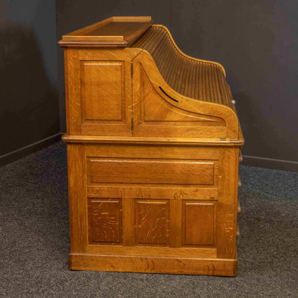A large Edwardian golden oak roll-top desk featuring a slatted tambour cover, an extensive fitted interior with small drawers and pigeonholes, and twin pedestals with carved wooden handles. View III