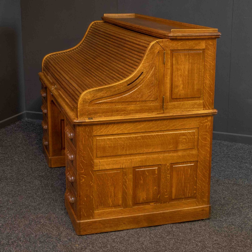 A large Edwardian golden oak roll-top desk featuring a slatted tambour cover, an extensive fitted interior with small drawers and pigeonholes, and twin pedestals with carved wooden handles. View VIII