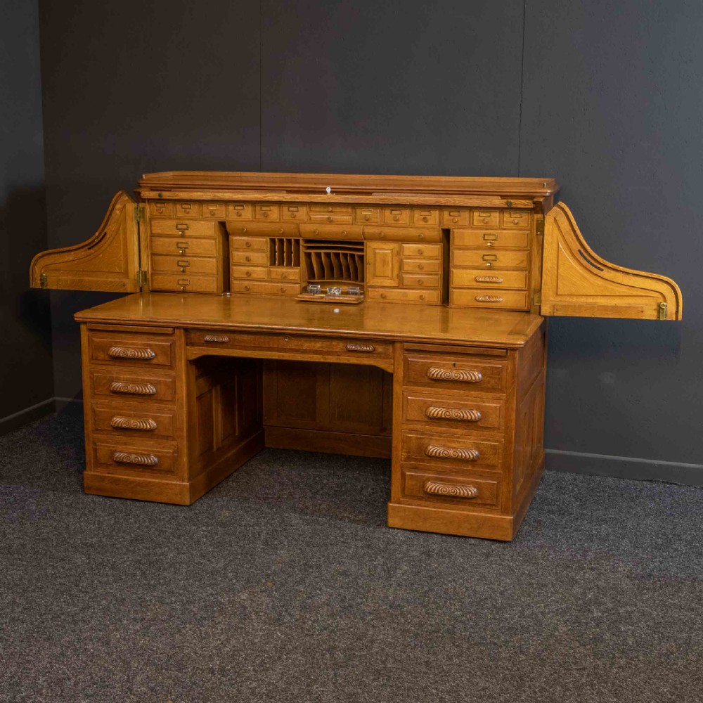 A large Edwardian golden oak roll-top desk featuring a slatted tambour cover, an extensive fitted interior with small drawers and pigeonholes, and twin pedestals with carved wooden handles. View X