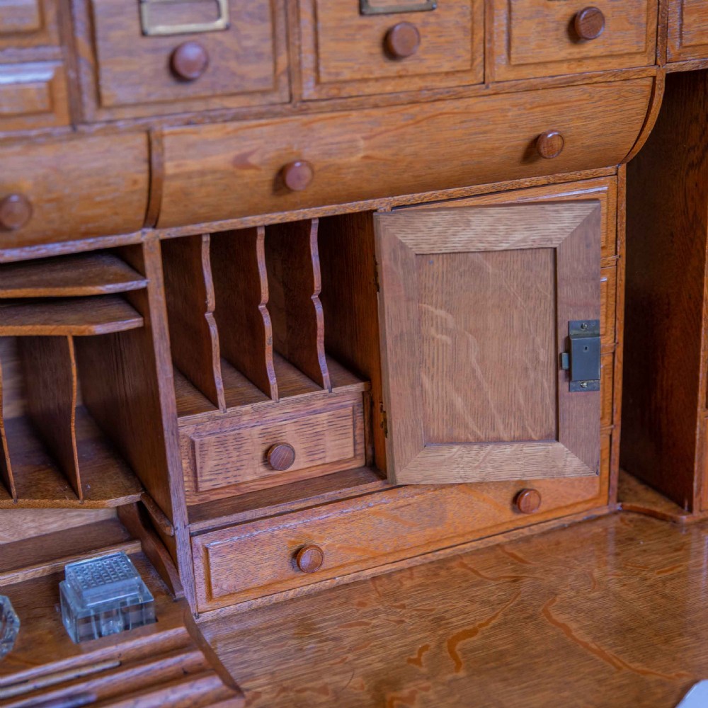 A large Edwardian golden oak roll-top desk featuring a slatted tambour cover, an extensive fitted interior with small drawers and pigeonholes, and twin pedestals with carved wooden handles. View XVI
