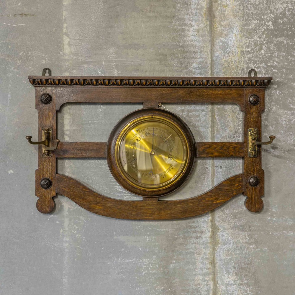 A wall-mounted carved oak coat rack featuring a central round brass aneroid barometer flanked by two double brass hooks.