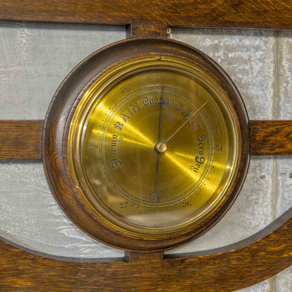 A wall-mounted carved oak coat rack featuring a central round brass aneroid barometer flanked by two double brass hooks. View II