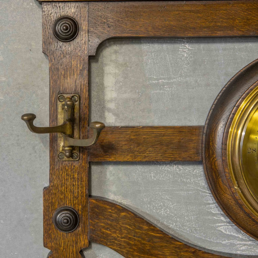 A wall-mounted carved oak coat rack featuring a central round brass aneroid barometer flanked by two double brass hooks. View III