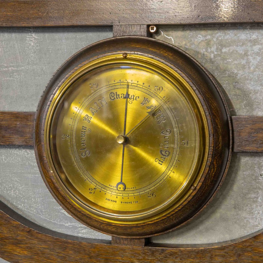 A wall-mounted carved oak coat rack featuring a central round brass aneroid barometer flanked by two double brass hooks. View V