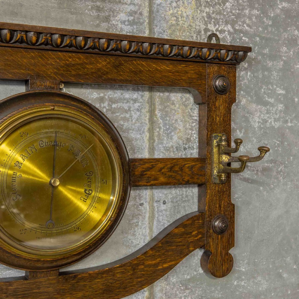 A wall-mounted carved oak coat rack featuring a central round brass aneroid barometer flanked by two double brass hooks. View VI
