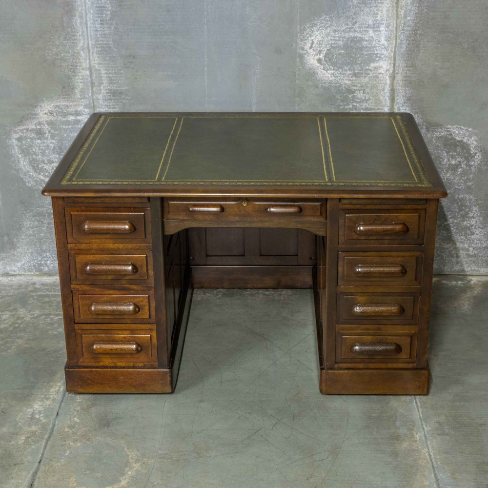 An Edwardian walnut pedestal desk featuring a green gold-tooled leather writing surface, nine drawers with integrated wooden handles, and a plinth base.