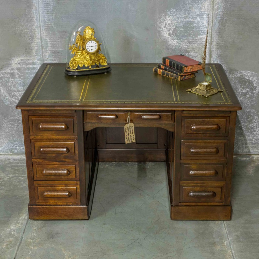 An Edwardian walnut pedestal desk featuring a green gold-tooled leather writing surface, nine drawers with integrated wooden handles, and a plinth base. View II