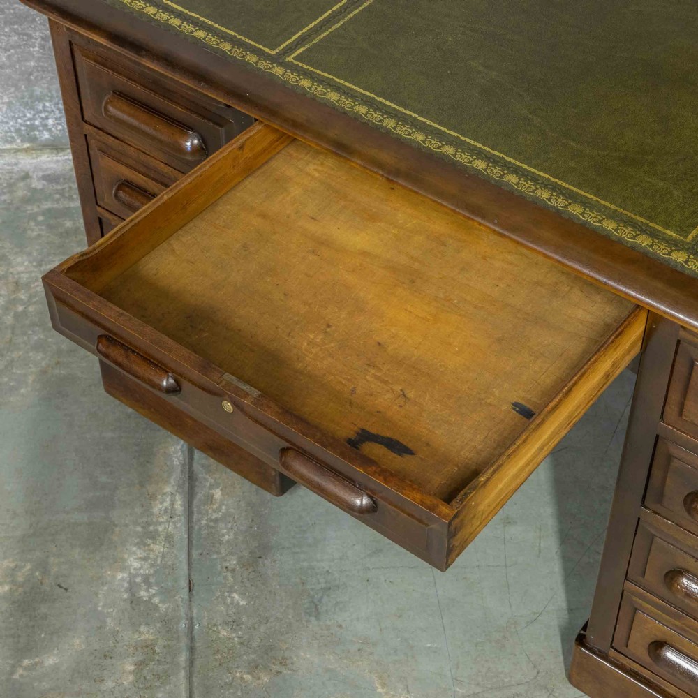 An Edwardian walnut pedestal desk featuring a green gold-tooled leather writing surface, nine drawers with integrated wooden handles, and a plinth base. View IV