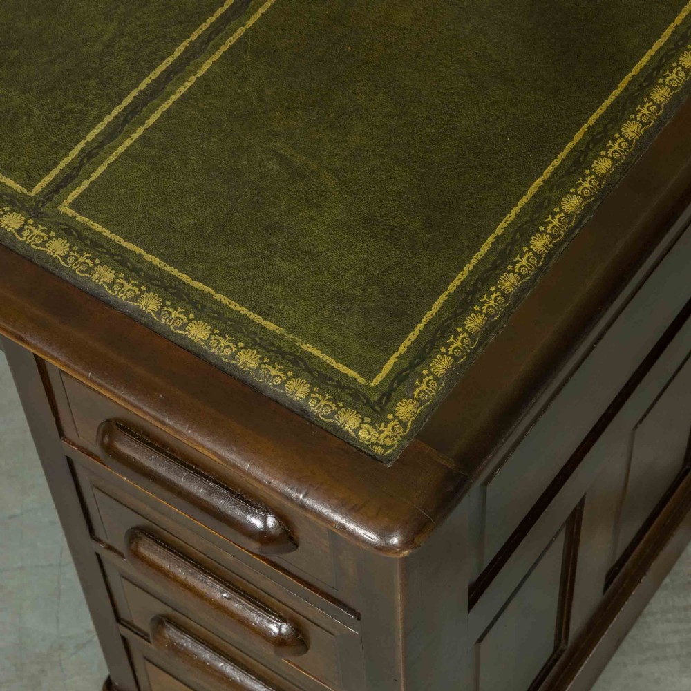 An Edwardian walnut pedestal desk featuring a green gold-tooled leather writing surface, nine drawers with integrated wooden handles, and a plinth base. View V