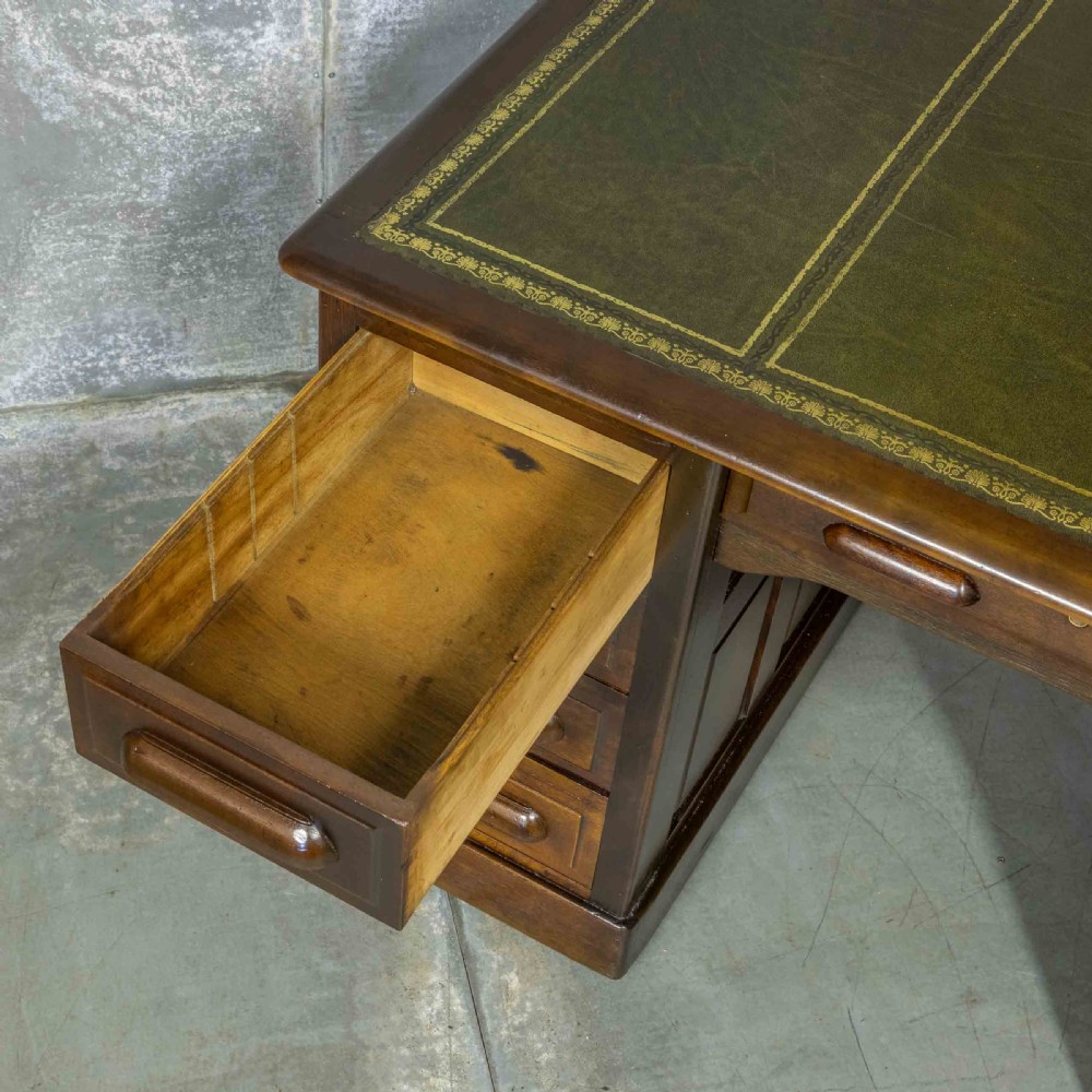 An Edwardian walnut pedestal desk featuring a green gold-tooled leather writing surface, nine drawers with integrated wooden handles, and a plinth base. View IX