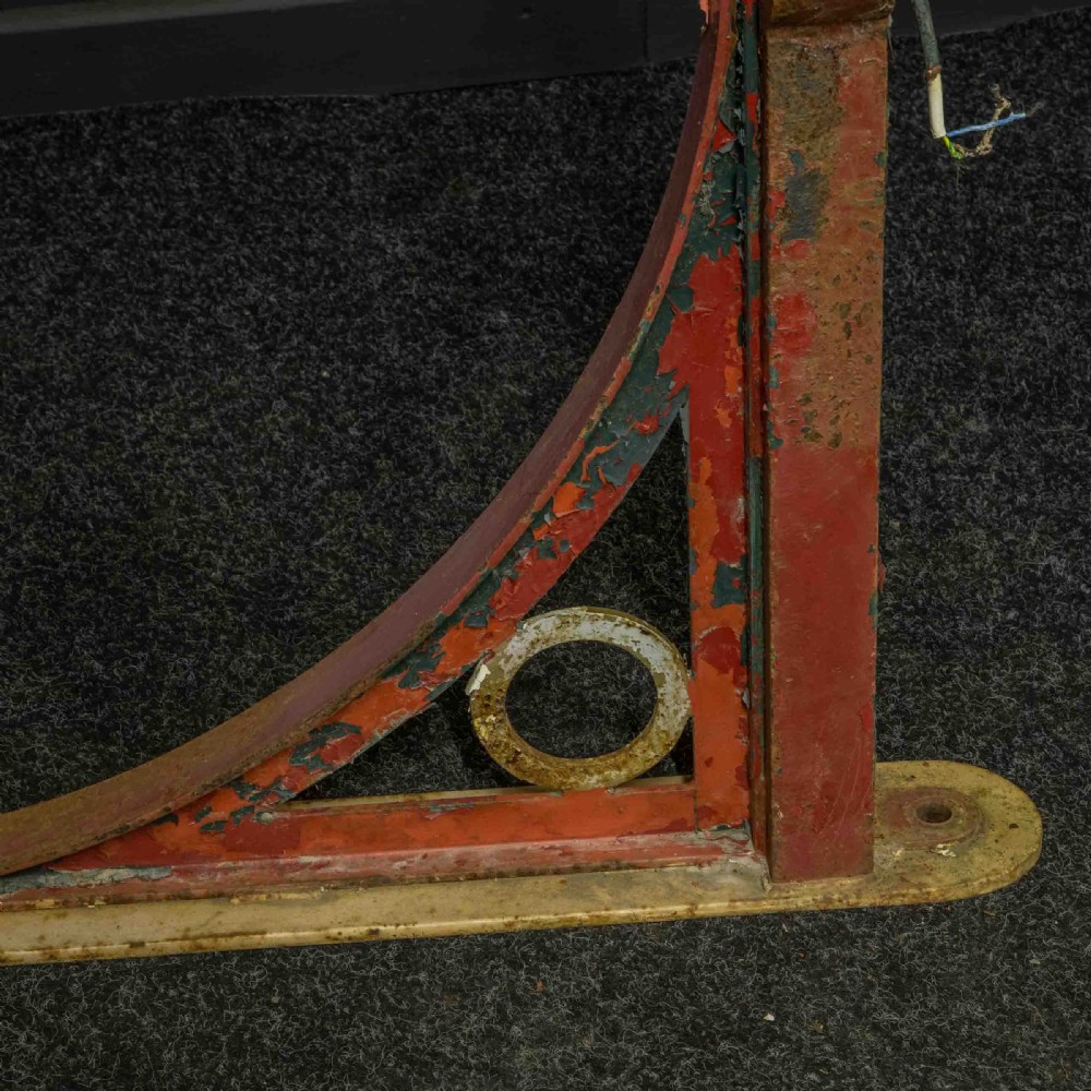 A heavy red painted cast iron wall bracket with a curved support bar and circular motif, showing distressed paint and metal details. View IX