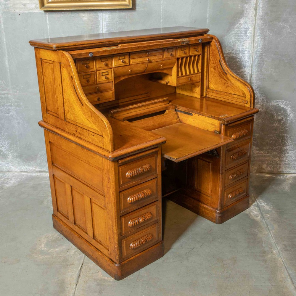 An Edwardian golden oak roll top desk featuring a flexible tambour shutter, fitted interior drawers, slotted pigeonholes, and twin pedestal bases with carved cup handles.
