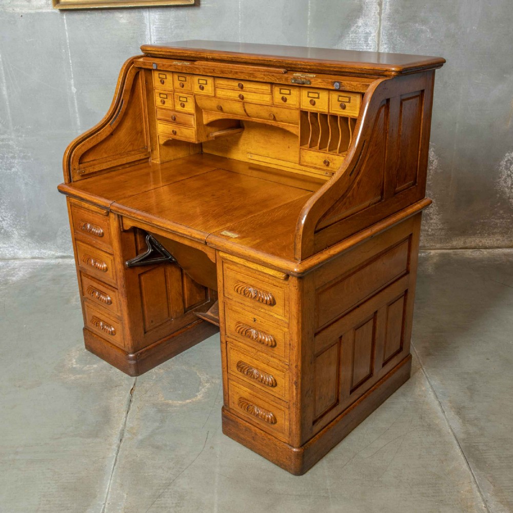 An Edwardian golden oak roll top desk featuring a flexible tambour shutter, fitted interior drawers, slotted pigeonholes, and twin pedestal bases with carved cup handles. View IV