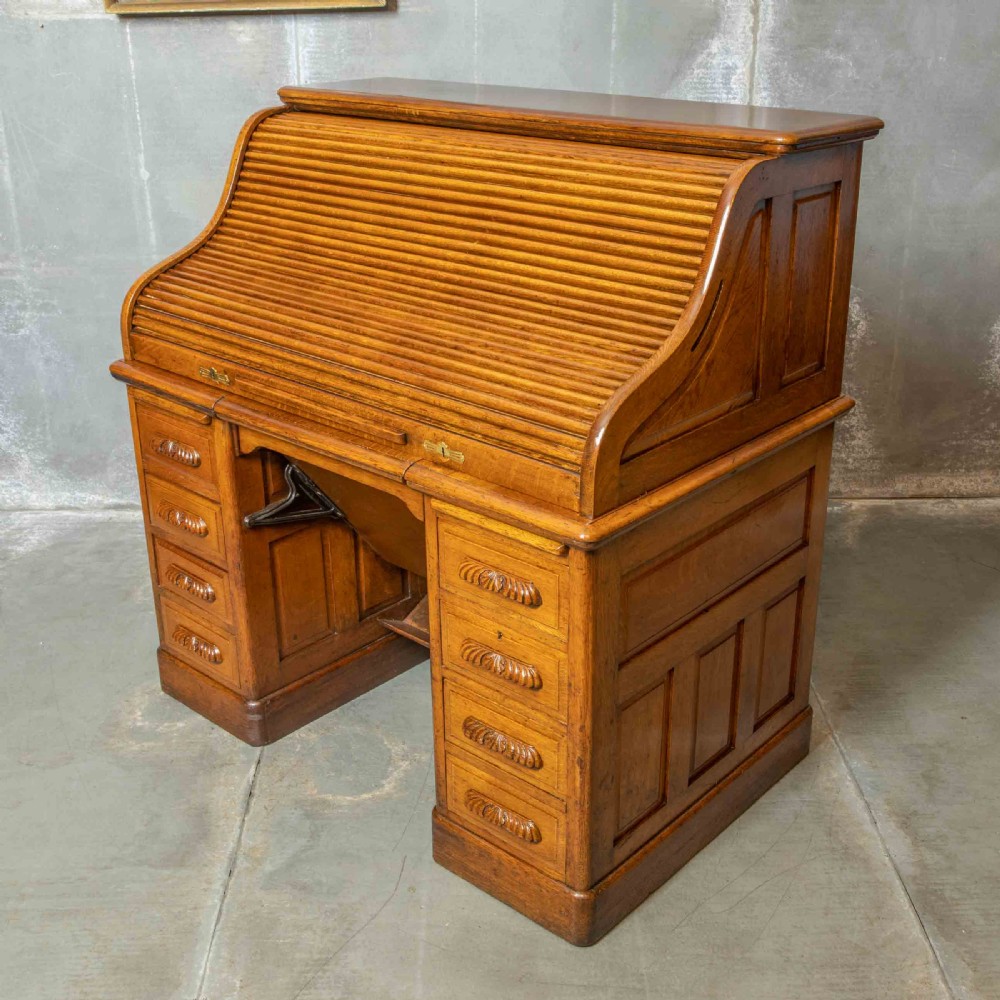 An Edwardian golden oak roll top desk featuring a flexible tambour shutter, fitted interior drawers, slotted pigeonholes, and twin pedestal bases with carved cup handles. View V