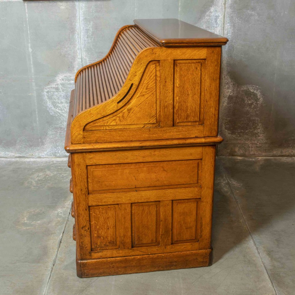 An Edwardian golden oak roll top desk featuring a flexible tambour shutter, fitted interior drawers, slotted pigeonholes, and twin pedestal bases with carved cup handles. View VII