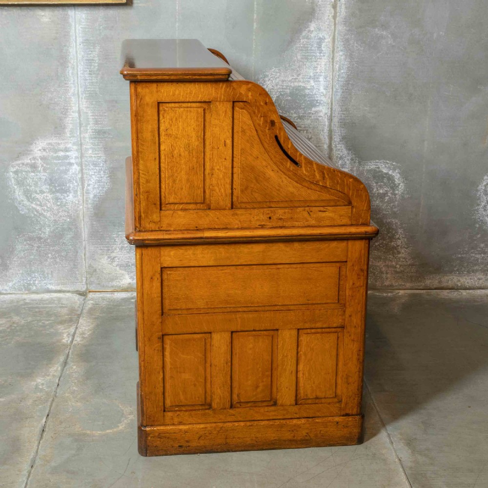 An Edwardian golden oak roll top desk featuring a flexible tambour shutter, fitted interior drawers, slotted pigeonholes, and twin pedestal bases with carved cup handles. View VIII