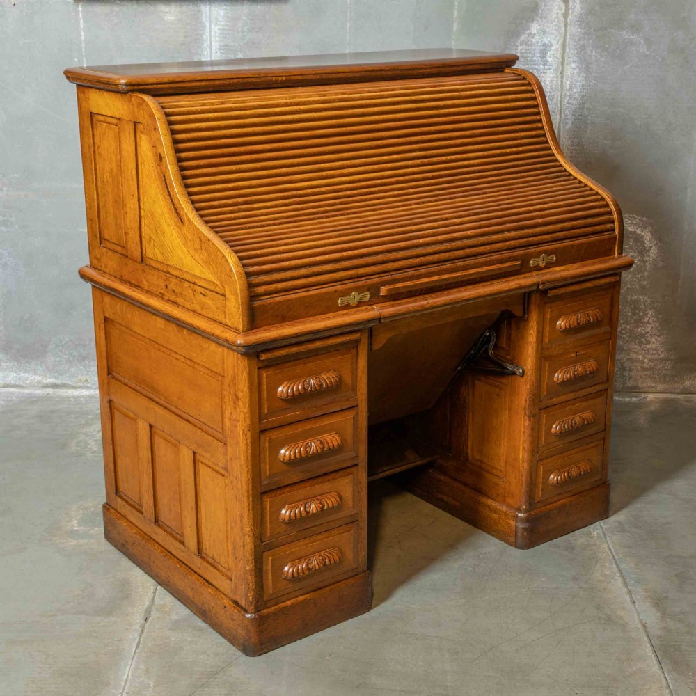 An Edwardian golden oak roll top desk featuring a flexible tambour shutter, fitted interior drawers, slotted pigeonholes, and twin pedestal bases with carved cup handles. View IX