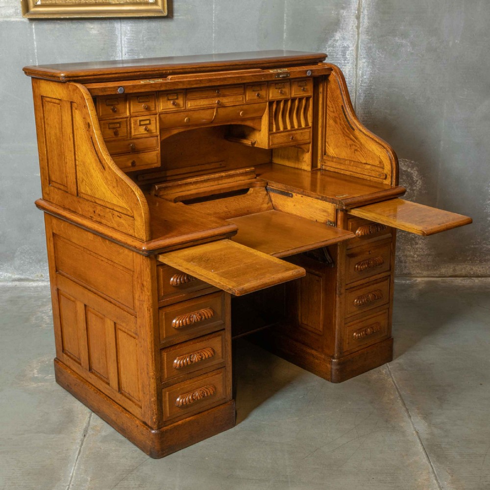 An Edwardian golden oak roll top desk featuring a flexible tambour shutter, fitted interior drawers, slotted pigeonholes, and twin pedestal bases with carved cup handles. View X