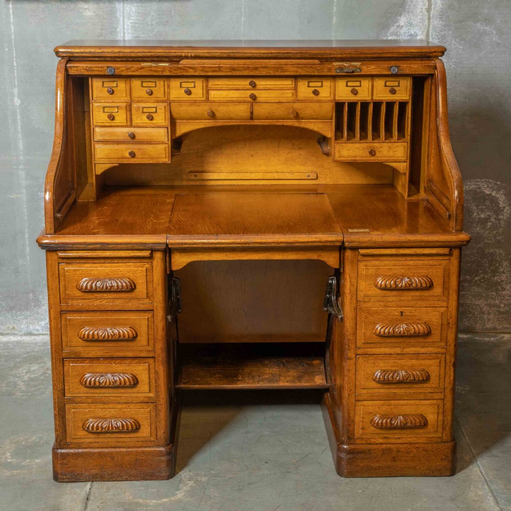 An Edwardian golden oak roll top desk featuring a flexible tambour shutter, fitted interior drawers, slotted pigeonholes, and twin pedestal bases with carved cup handles. View XI