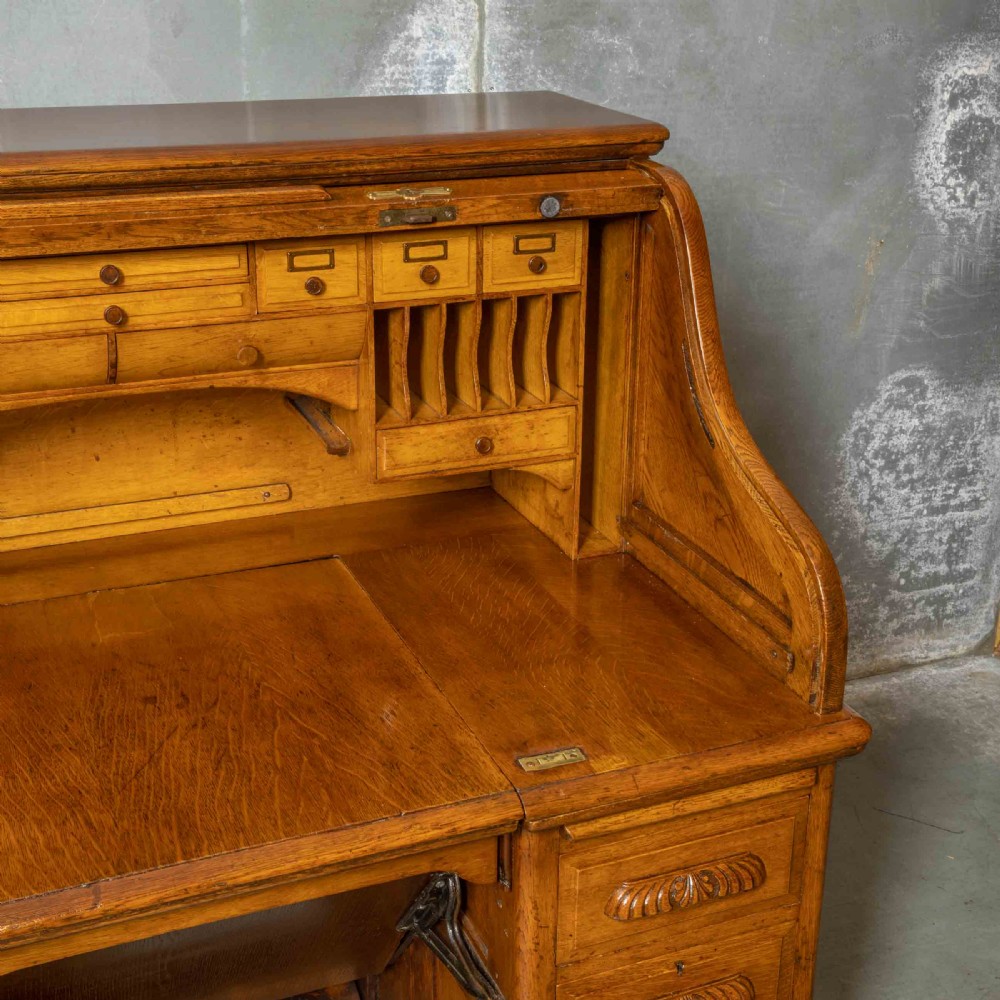 An Edwardian golden oak roll top desk featuring a flexible tambour shutter, fitted interior drawers, slotted pigeonholes, and twin pedestal bases with carved cup handles. View XII