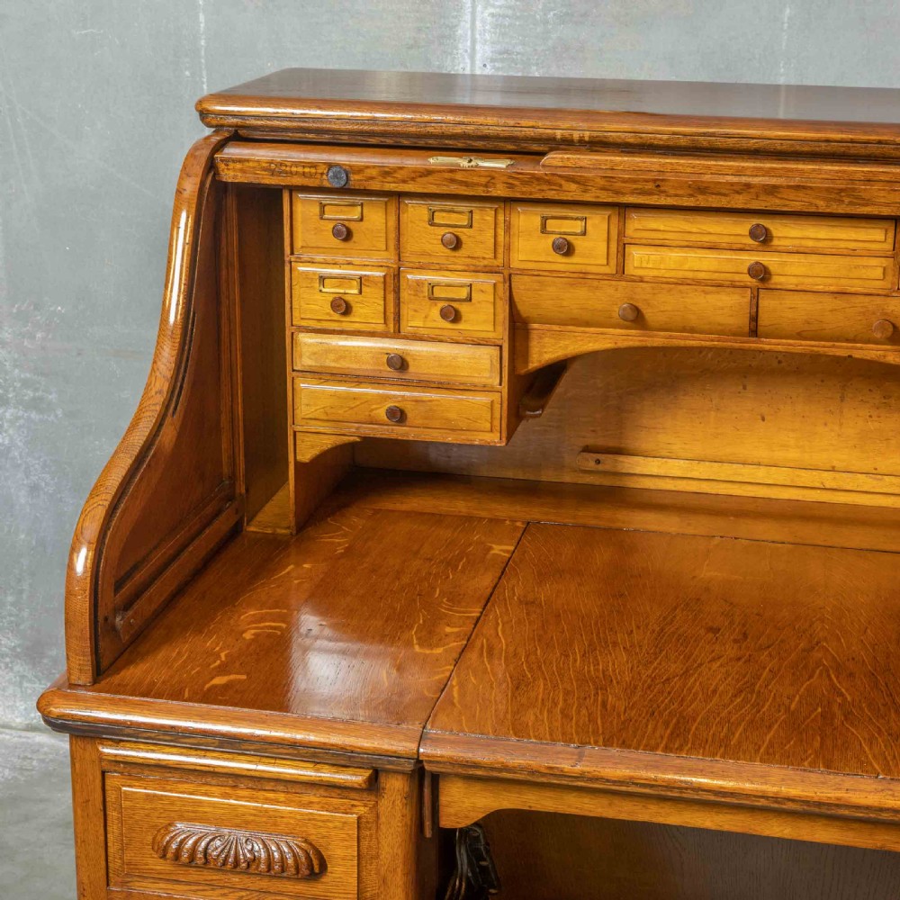 An Edwardian golden oak roll top desk featuring a flexible tambour shutter, fitted interior drawers, slotted pigeonholes, and twin pedestal bases with carved cup handles. View XIV