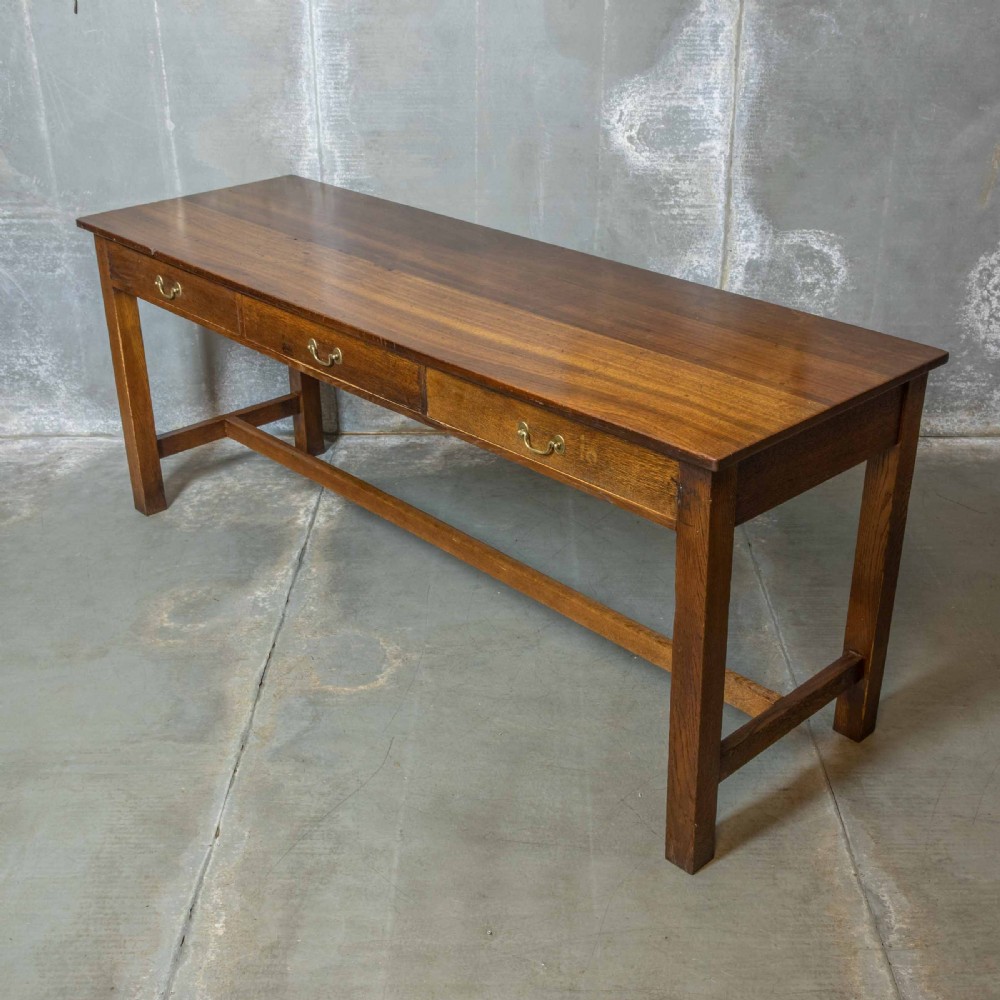 A long, tall oak refectory table featuring three frieze drawers with brass handles, straight legs, and a low H-stretcher base.