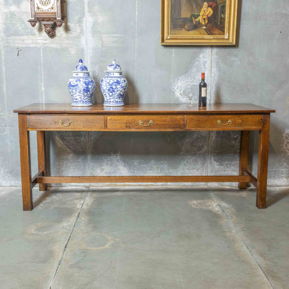A long, tall oak refectory table featuring three frieze drawers with brass handles, straight legs, and a low H-stretcher base. View II