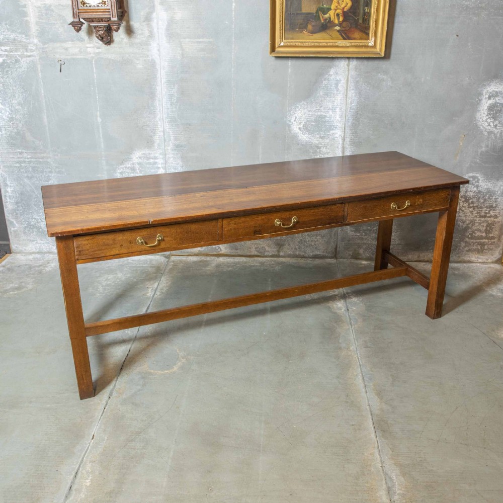 A long, tall oak refectory table featuring three frieze drawers with brass handles, straight legs, and a low H-stretcher base. View X