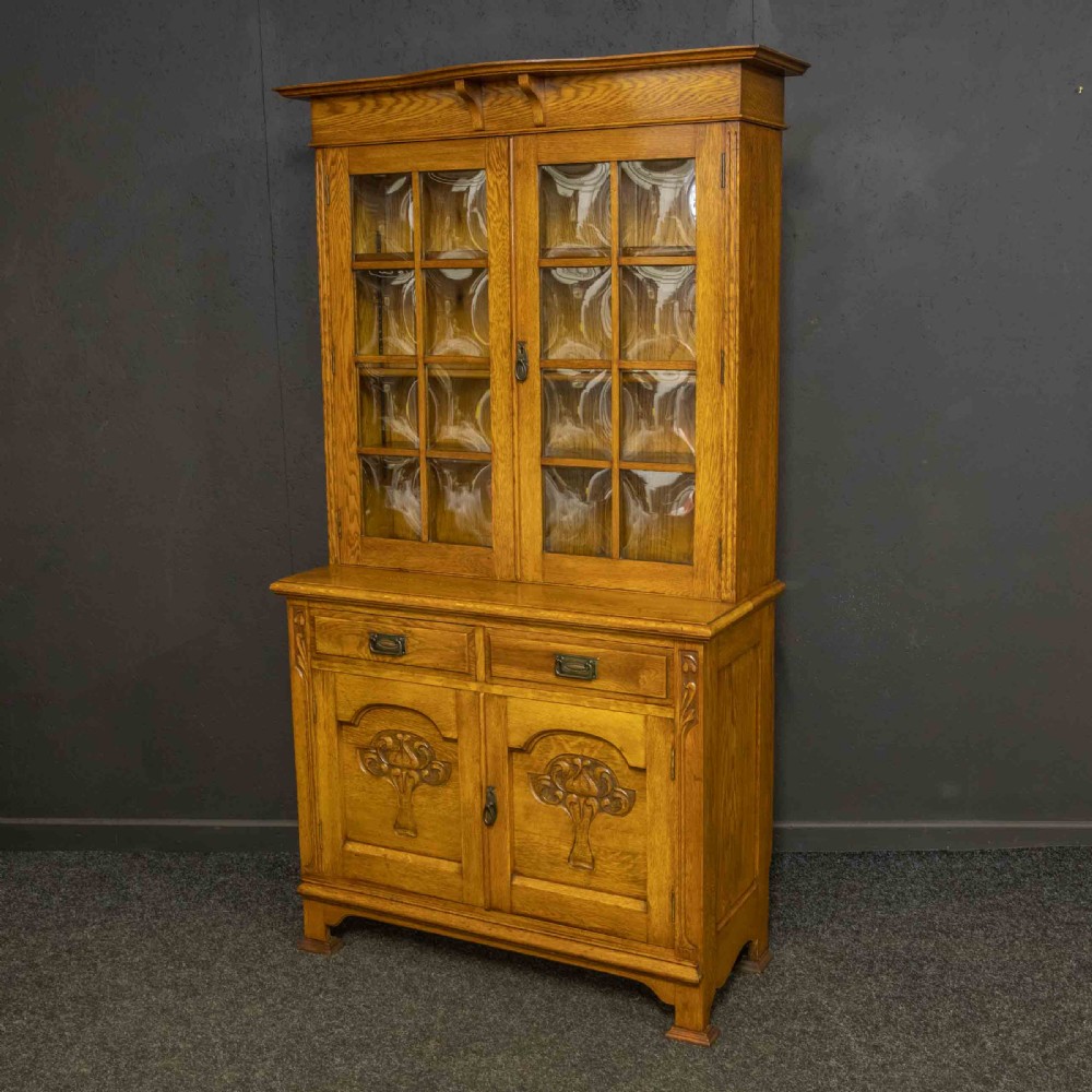 An Art Nouveau golden oak glazed bookcase featuring upper glass door panels and lower carved doors with stylised floral motifs.