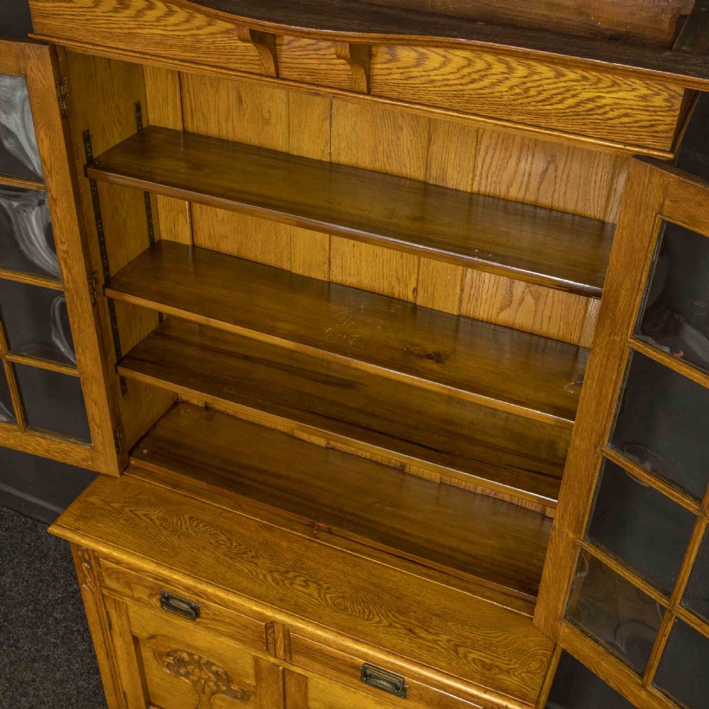 An Art Nouveau golden oak glazed bookcase featuring upper glass door panels and lower carved doors with stylised floral motifs. View II