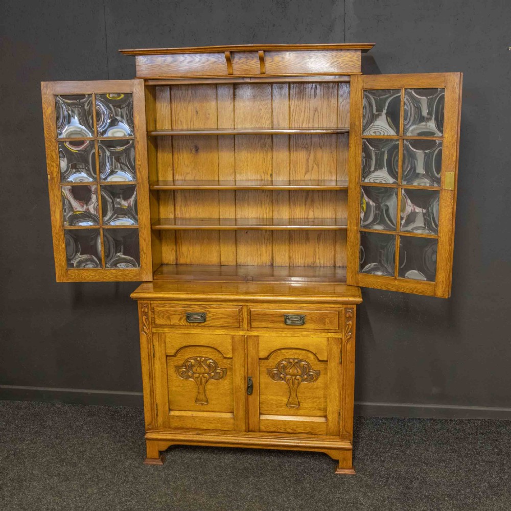 An Art Nouveau golden oak glazed bookcase featuring upper glass door panels and lower carved doors with stylised floral motifs. View V