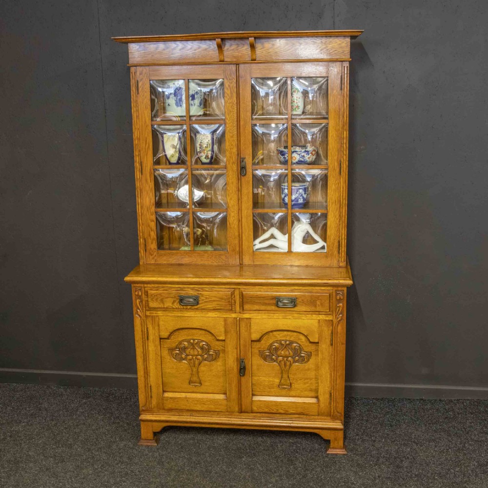 An Art Nouveau golden oak glazed bookcase featuring upper glass door panels and lower carved doors with stylised floral motifs. View VI