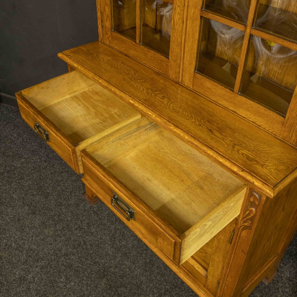 An Art Nouveau golden oak glazed bookcase featuring upper glass door panels and lower carved doors with stylised floral motifs. View VII