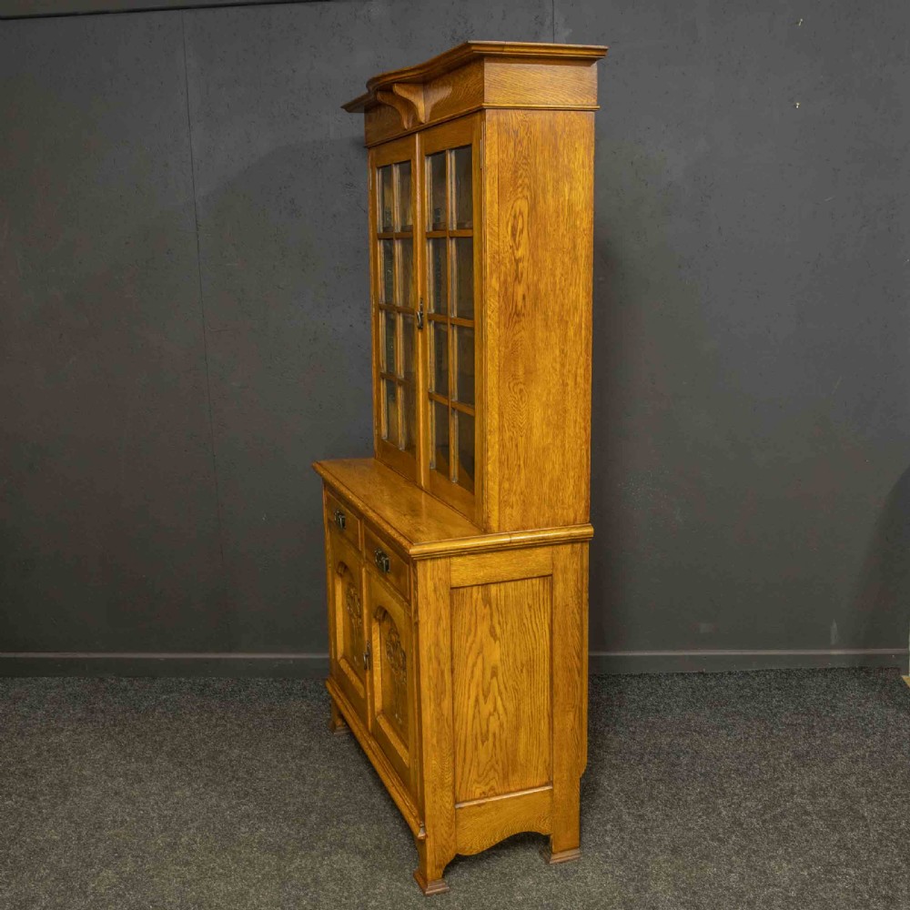An Art Nouveau golden oak glazed bookcase featuring upper glass door panels and lower carved doors with stylised floral motifs. View VIII