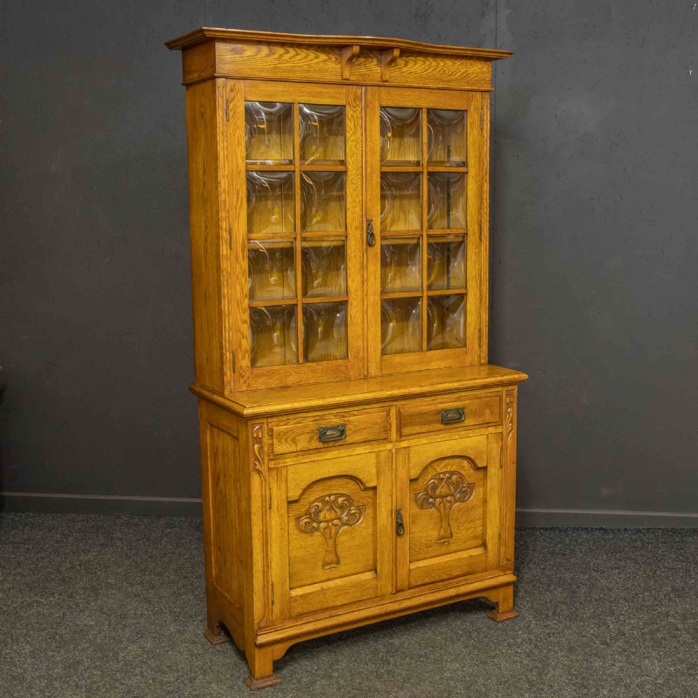 An Art Nouveau golden oak glazed bookcase featuring upper glass door panels and lower carved doors with stylised floral motifs. View XI