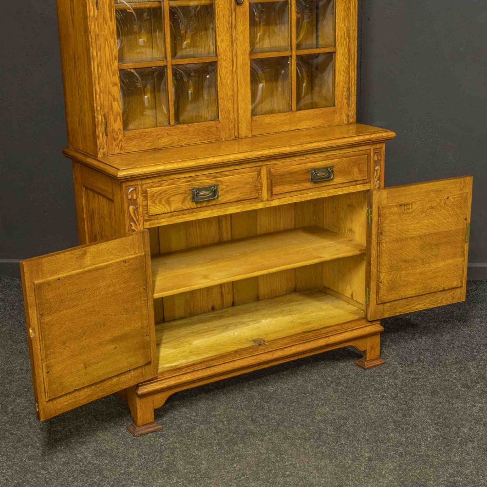 An Art Nouveau golden oak glazed bookcase featuring upper glass door panels and lower carved doors with stylised floral motifs. View XII