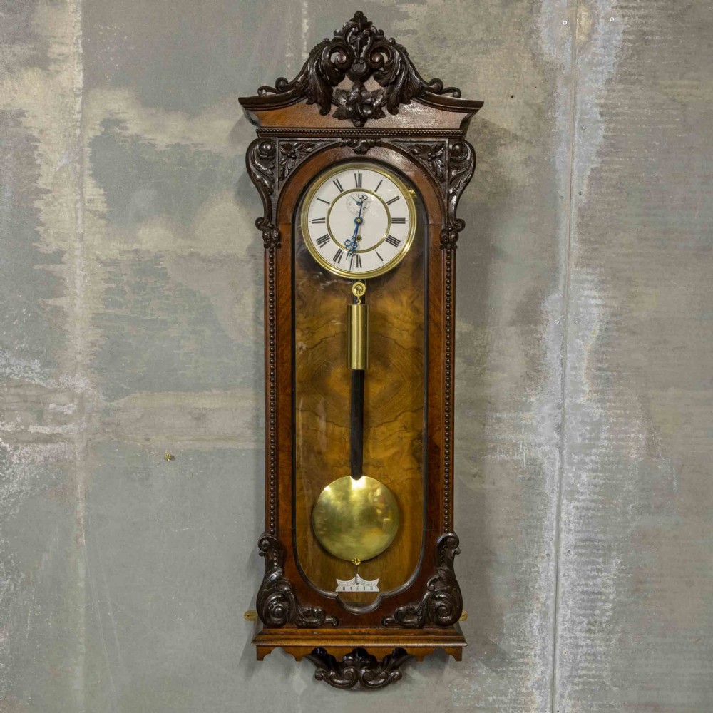A Victorian Vienna regulator wall clock featuring a dark walnut carved timber case, enamel dial, and a visible brass pendulum behind a glass door.