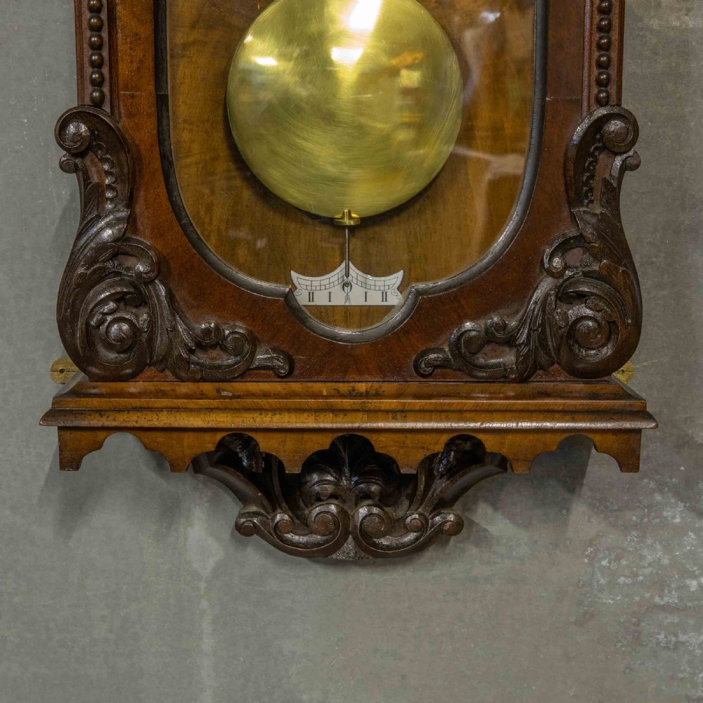 A Victorian Vienna regulator wall clock featuring a dark walnut carved timber case, enamel dial, and a visible brass pendulum behind a glass door. View II