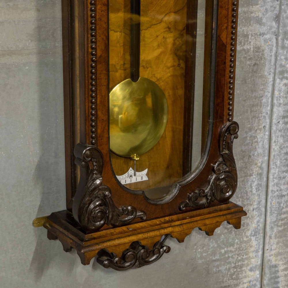 A Victorian Vienna regulator wall clock featuring a dark walnut carved timber case, enamel dial, and a visible brass pendulum behind a glass door. View III