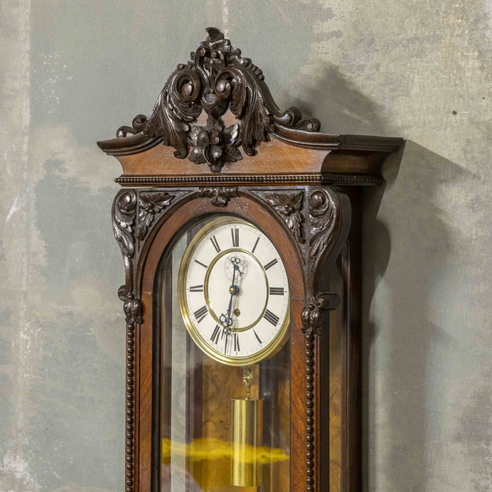 A Victorian Vienna regulator wall clock featuring a dark walnut carved timber case, enamel dial, and a visible brass pendulum behind a glass door. View IV