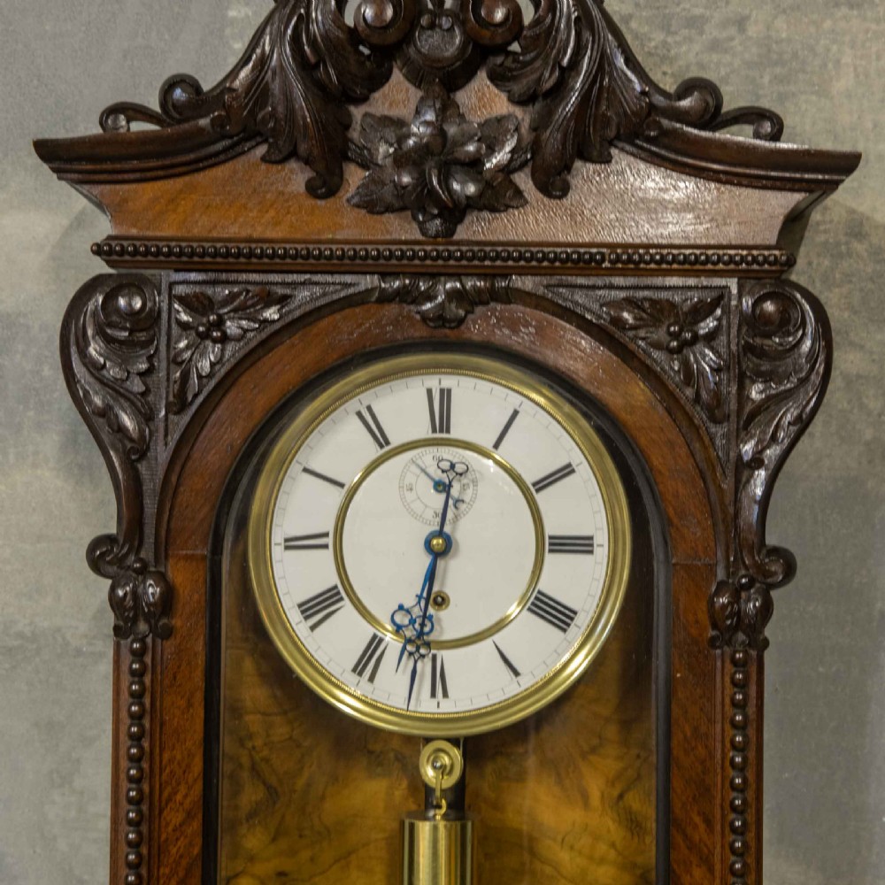 A Victorian Vienna regulator wall clock featuring a dark walnut carved timber case, enamel dial, and a visible brass pendulum behind a glass door. View V