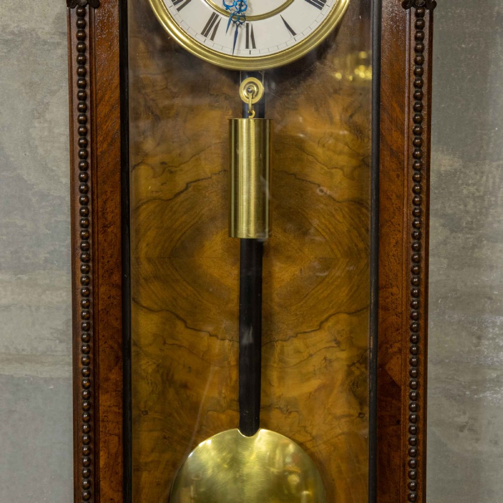 A Victorian Vienna regulator wall clock featuring a dark walnut carved timber case, enamel dial, and a visible brass pendulum behind a glass door. View VI