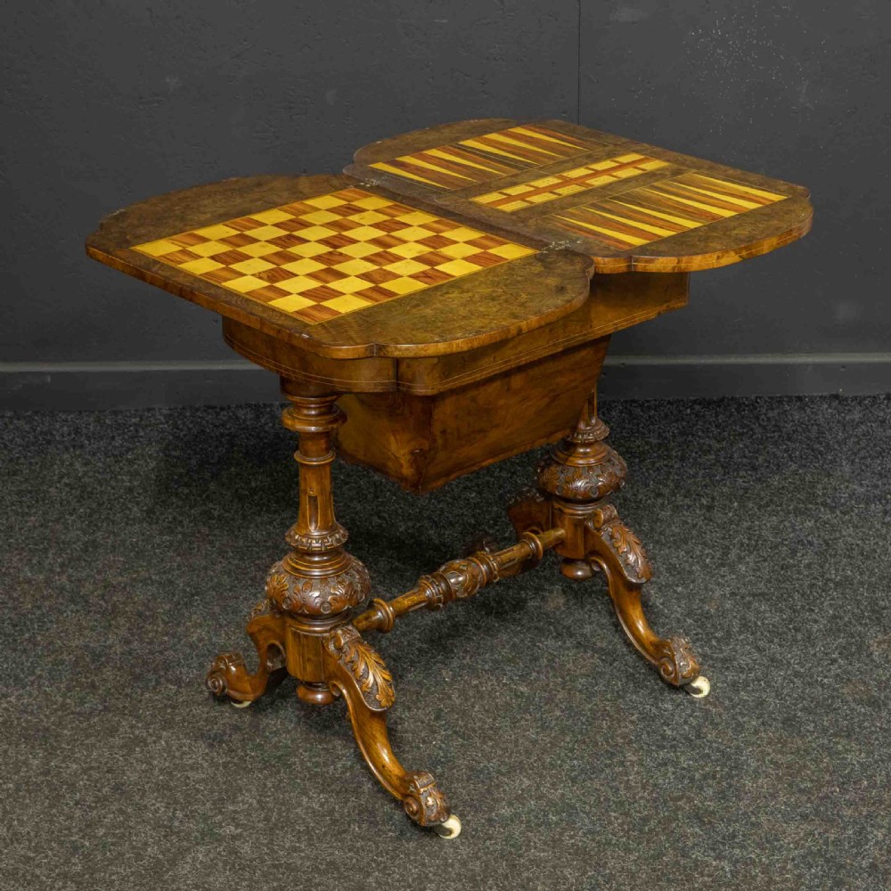 A Victorian burr walnut fold-over games and sewing table displaying an inlaid chess and backgammon board supported by carved pedestals, a stretcher base, and castors.