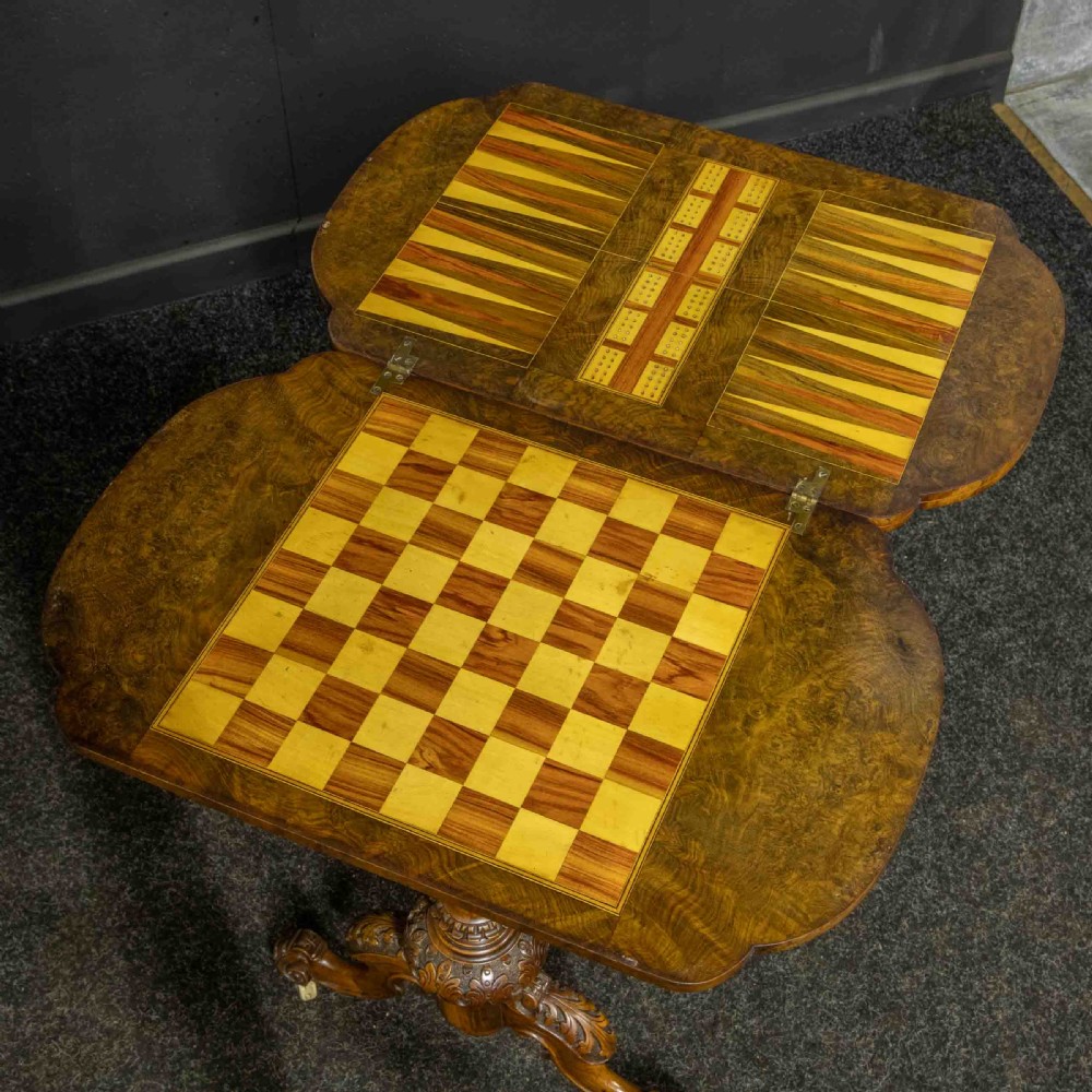 A Victorian burr walnut fold-over games and sewing table displaying an inlaid chess and backgammon board supported by carved pedestals, a stretcher base, and castors. View V