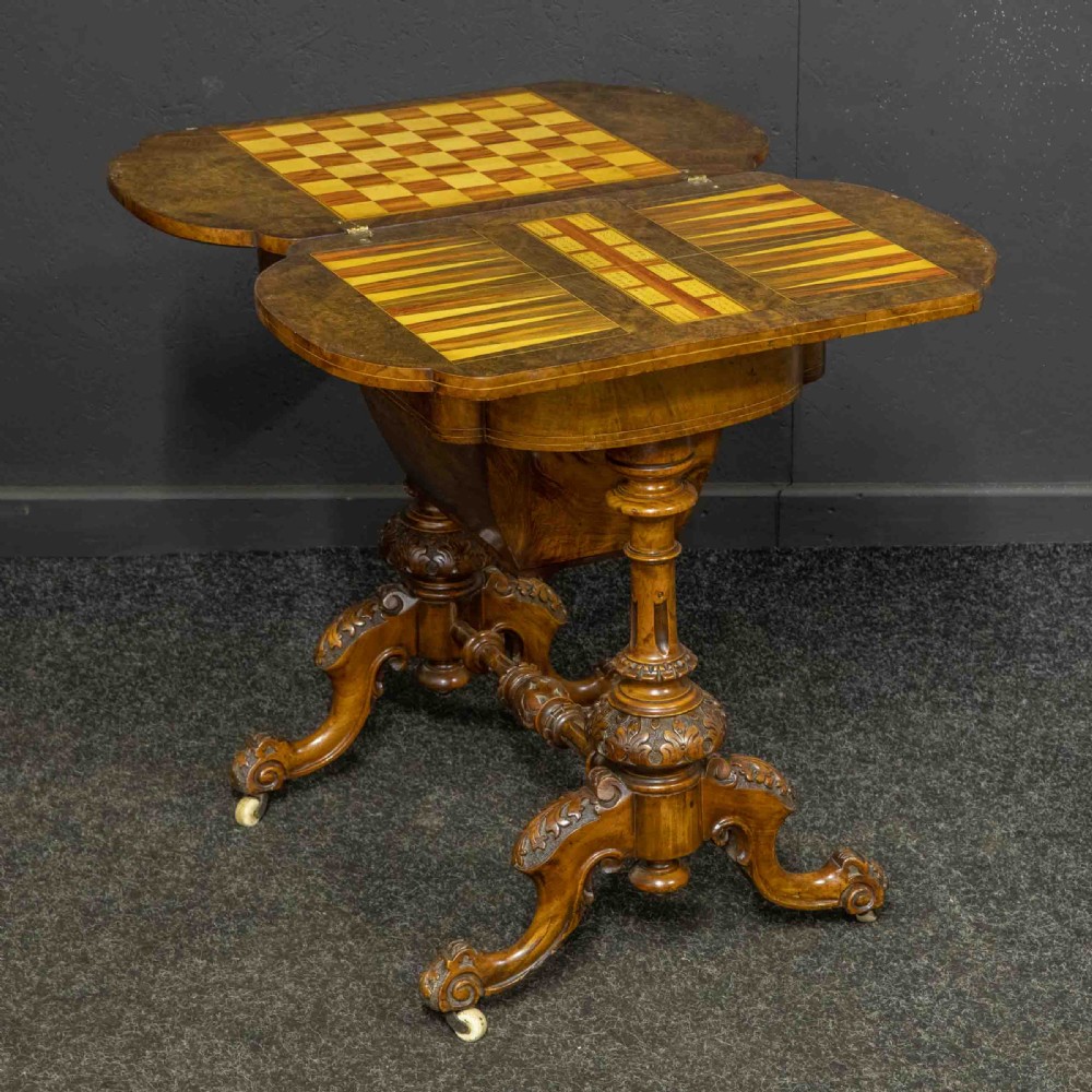 A Victorian burr walnut fold-over games and sewing table displaying an inlaid chess and backgammon board supported by carved pedestals, a stretcher base, and castors. View VI