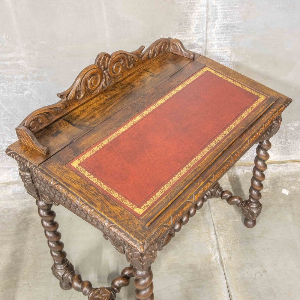 A Victorian carved oak writing desk featuring a sloped red leather surface, barley twist legs, and carved gallery details. View VI