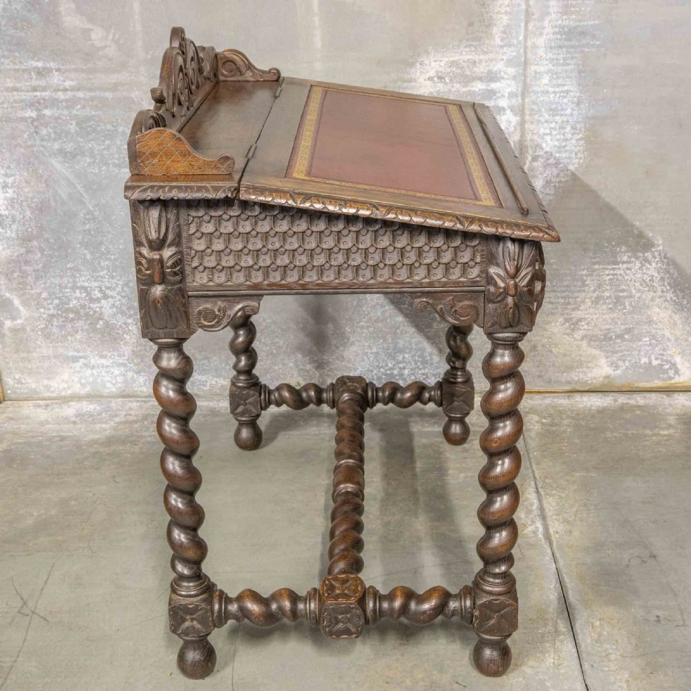 A Victorian carved oak writing desk featuring a sloped red leather surface, barley twist legs, and carved gallery details. View VII