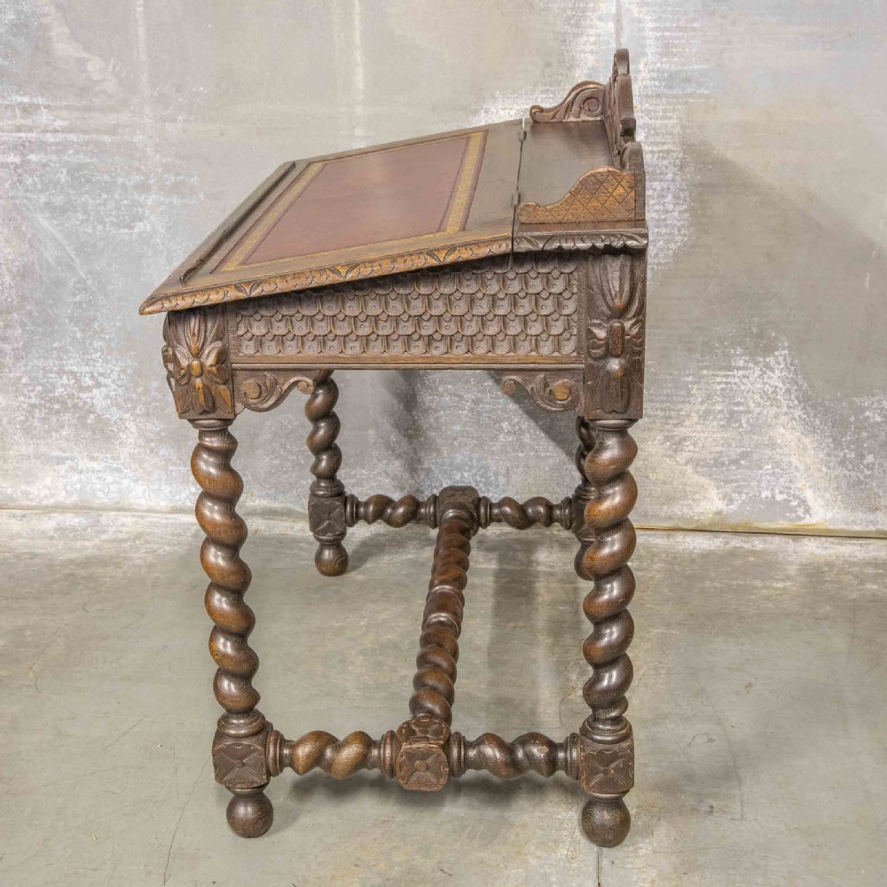 A Victorian carved oak writing desk featuring a sloped red leather surface, barley twist legs, and carved gallery details. View IX